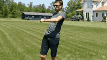 a man in a gray shirt and black shorts is standing in a grassy field in front of a house