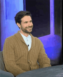 a man with a beard is sitting at a table wearing a brown sweater and smiling .