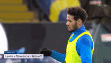 a man in a blue and yellow vest stands on a soccer field in front of a banner that says coming next