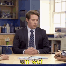 a man in a suit and tie is sitting at a table in a classroom talking to a group of children .