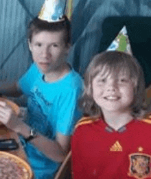 a boy and a girl are sitting at a table with pizza and wearing party hats .