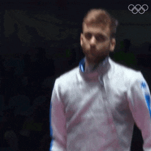 a man in a white jacket is standing in front of a black background with the olympic rings in the background .