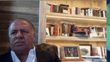 a man in a suit is sitting in front of a bookshelf filled with books and framed pictures .