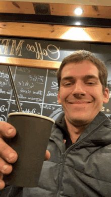 a man holding a cup of coffee in front of a coffee menu board