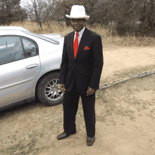 a man in a suit and white hat stands in front of a car