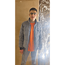 a young man wearing a grey jacket and an orange t-shirt stands in front of a marble wall