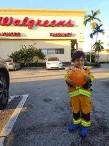 a little boy dressed as a fireman holds a pumpkin in front of a walgreens liquors and pharmacy