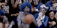 a man with blue paint on his face is holding a basketball in front of a crowd of people wearing duke shirts