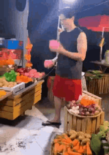 a man in a black tank top and red shorts is standing in front of a fruit and vegetable stand .