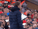 a man in a blue jacket stands in front of a crowd with a scoreboard that says bre on it