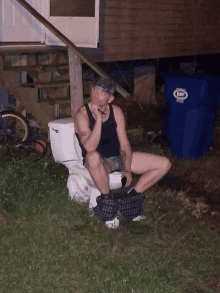 a man sits on a toilet next to a blue bin that says ewp