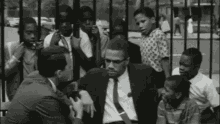 a black and white photo of a man in a suit and tie sitting in front of a fence surrounded by children .