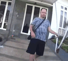 a man in a tie dye shirt and black shorts is standing in front of a house holding a mop .