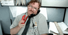 a man in a cubicle talking on a phone with the word bill written on his shirt