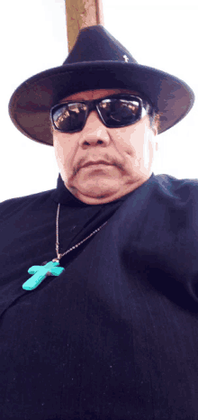 a man wearing a black hat and sunglasses has a green cross necklace around his neck