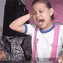 a little girl wearing pink suspenders and a white shirt is sitting at a table with her mouth open .