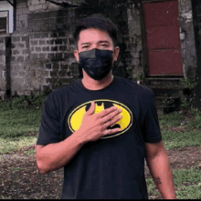 a man wearing a mask and a batman t-shirt is standing in front of a brick wall .