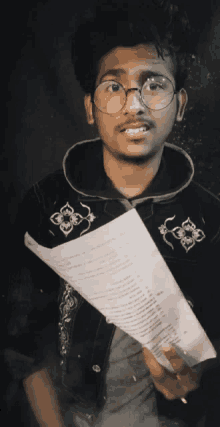 a young man wearing glasses and a black jacket holds a piece of paper