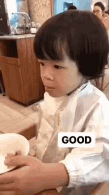 a little boy is sitting at a table with a bowl of food and a sign that says good on it .