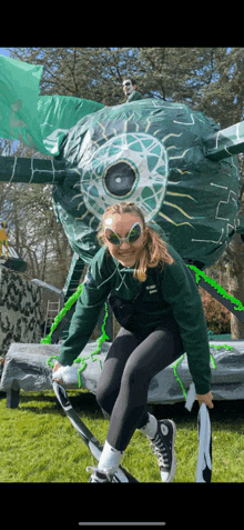 a girl wearing sunglasses and a green jacket is standing in front of a green inflatable object .