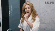 a woman in a bathrobe is brushing her teeth in front of a mirror that says bazaar on it