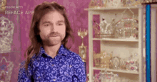 a man with long hair and a beard wearing a blue shirt is standing in front of a shelf full of crowns .