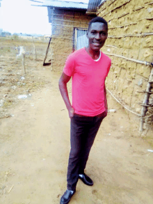 a young man wearing a red shirt and black pants stands in front of a stone wall