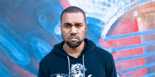 kanye west is wearing a black hoodie and standing in front of a blue and red wall .