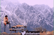 a man sits on a picnic table in front of a mountain with a sign that says ' bhebatsam ' on it