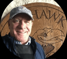 a man wearing a hat stands in front of a wooden sign that says hawk