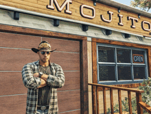 a man in a cowboy hat and plaid shirt stands in front of a mojito restaurant