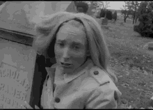 a woman in a trench coat is standing in front of a grave in a cemetery with her hair blowing in the wind .