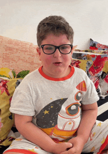 a young boy wearing glasses and a shirt with a rocket on it sits on a bed