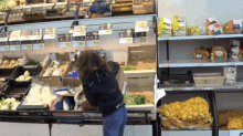 a woman standing in front of a display of fruits and vegetables with a bag of potatoes on the bottom shelf