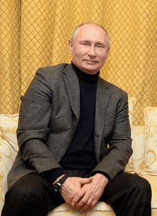 a man in a suit and turtleneck sits on a couch with his hands folded