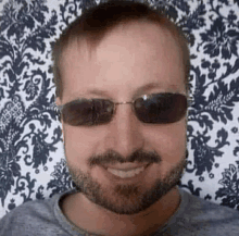 a man with a beard and sunglasses is smiling in front of a patterned wallpaper .