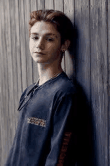 a young man is leaning against a wooden wall and wearing a blue shirt .