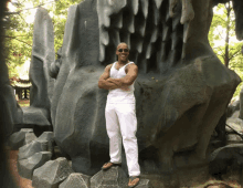 a man in white pants and a white tank top stands in front of a large rock