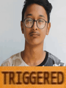 a young man wearing glasses and a camouflage shirt is behind a yellow sign that says triggered