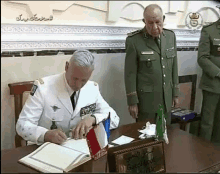 a man in a military uniform is signing a guestbook while another man looks on .