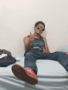 a man wearing sunglasses gives a thumbs up while sitting on a bed