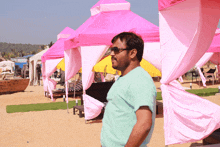 a man wearing sunglasses stands in front of pink canopies on the beach