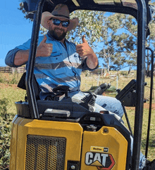 a man wearing a cowboy hat is giving a thumbs up while sitting in a yellow cat tractor