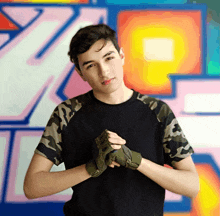 a young man wearing gloves and a black shirt stands in front of a wall with graffiti on it