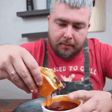 a man wearing a red shirt that says called to grill is dipping a piece of food into a bowl of sauce