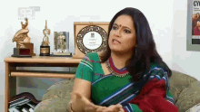 a woman in a green and red striped saree is sitting on a couch in front of a table with trophies on it