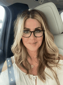 a woman wearing glasses and a necklace smiles while sitting in a car