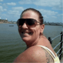 a woman wearing sunglasses and a tattoo on her arm smiles in front of a body of water