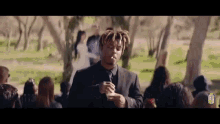 a man in a suit is smoking a cigarette while standing in front of a crowd of people at a funeral .