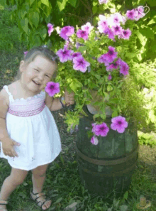 a little girl in a white dress is standing next to a potted plant with purple flowers